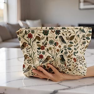 Peut inclure: Une pochette rectangulaire à fermeture éclair avec un fond crème orné d'un motif répétitif de petits oiseaux, de fleurs rouges et de feuillage vert. Le tissu présente une texture finement côtelée et une fermeture éclair en métal doré.