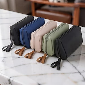 Puede incluir: Cinco estuches rectangulares con cremallera dispuestos en fila, en colores negro, azul marino, beige, verde salvia y gris carbón. Cada estuche incluye una lengüeta de tela a juego unida a un cierre de cremallera de metal plateado.
