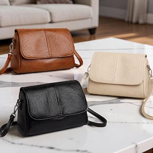 May include: Three rectangular crossbody bags with a textured finish and a front flap closure. The bags are available in solid brown, black, and cream colors, each featuring a matching adjustable shoulder strap attached with silver-toned metal hardware.