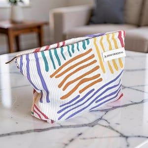 Può includere: Una pochette bianca con cerniera, caratterizzata da una consistenza a coste e un motivo astratto colorato con linee dipinte in viola, verde acqua, arancione e giallo. Un'etichetta rettangolare bianca in tessuto con il testo nero A GOGO ORIGINAL è cucita sul lato.