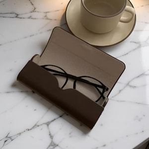 Puede incluir: Un estuche para gafas rectangular de color marrón oscuro con un forro interior beige, mostrado en posición abierta. Dentro del estuche descansa un par de gafas con montura negra. Una taza y un plato de cerámica beige están cerca del estuche.