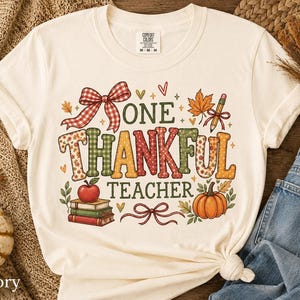 Puede incluir: Una camiseta de manga corta color marfil con un gráfico que incluye el texto "ONE THANKFUL TEACHER" en letras coloridas y estampadas. El diseño presenta un lazo de cuadros rojos, una manzana roja sobre libros, un lápiz, hojas y una calabaza.