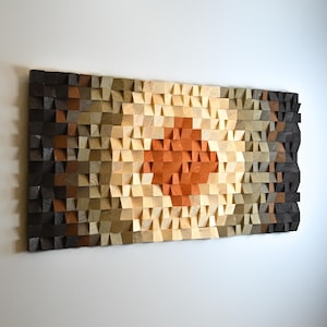 May include: A rectangular wall art piece composed of numerous small, individual wooden blocks of varying depths and shades. The colors transition from dark charcoal and brown at the edges to lighter tan and a central focal point of warm orange.