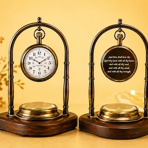 Puede incluir: Dos relojes de escritorio de latón de estilo antiguo con bases de madera oscura. Uno tiene una esfera blanca con números negros, el otro muestra un texto religioso grabado. Ambos cuelgan de marcos de latón arqueados sobre una brújula.