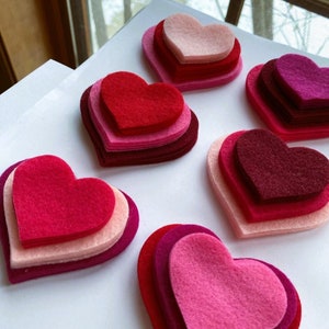 May include: Six layered felt hearts in various shades of pink and red. The hearts are arranged in a semi-circle on a white surface.