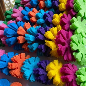 Op de afbeelding: Een verzameling van viltbloemvormen in verschillende kleuren, waaronder groen, roze, blauw, oranje, paars, geel en rood. De bloemen zijn in een cirkelvormig patroon gerangschikt, met de grootste bloemen in het midden en de kleinste bloemen aan de buitenkant. Er zijn ook verschillende kleine viltcirkel in verschillende kleuren.
