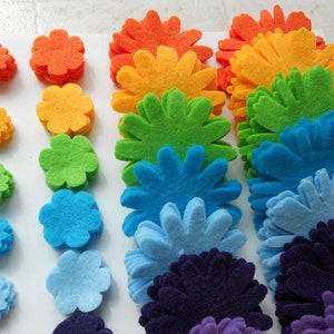K&ouml;nnte beinhalten: Ein Regenbogen aus Filzblumen, beginnend mit Orange und endend mit Lila. Jede Blume besteht aus mehreren Schichten Filz, wodurch ein dreidimensionaler Effekt entsteht.