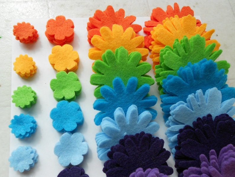 K&ouml;nnte beinhalten: Ein Regenbogen aus Filzblumen, beginnend mit Orange und endend mit Lila. Jede Blume besteht aus mehreren Schichten Filz, wodurch ein dreidimensionaler Effekt entsteht.