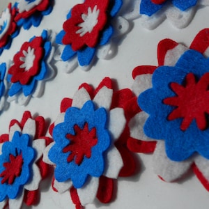 May include: A collection of felt flowers in red, white, and blue. The flowers are layered and have a starburst design in the center.