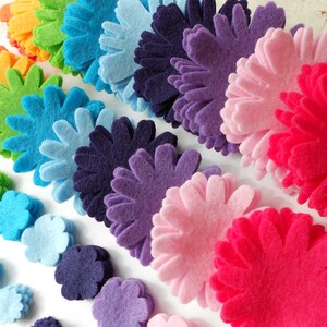 K&ouml;nnte beinhalten: Eine Regenbogen-Auswahl an Filzblumenformen in verschiedenen Gr&ouml;&szlig;en von klein bis gro&szlig;. Die Blumen sind in Reihen gestapelt, wobei die kleinsten Blumen unten und die gr&ouml;&szlig;ten Blumen oben liegen. Die Blumen sind alle aus Filz und haben eine Vielzahl von Farben, darunter Rot, Orange, Gelb, Gr&uuml;n, Blau, Lila und Pink.