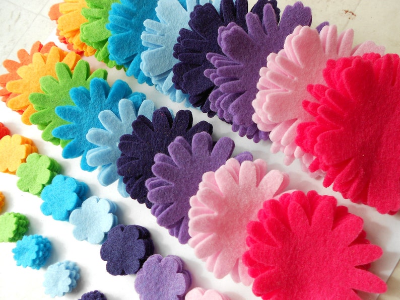 K&ouml;nnte beinhalten: Eine Regenbogen-Auswahl an Filzblumenformen in verschiedenen Gr&ouml;&szlig;en von klein bis gro&szlig;. Die Blumen sind in Reihen gestapelt, wobei die kleinsten Blumen unten und die gr&ouml;&szlig;ten Blumen oben liegen. Die Blumen sind alle aus Filz und haben eine Vielzahl von Farben, darunter Rot, Orange, Gelb, Gr&uuml;n, Blau, Lila und Pink.
