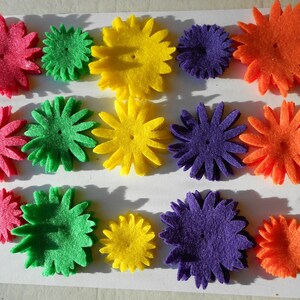 May include: A collection of felt flower embellishments in various colors, including red, green, yellow, purple, and orange. The flowers are layered and have a simple, symmetrical design.