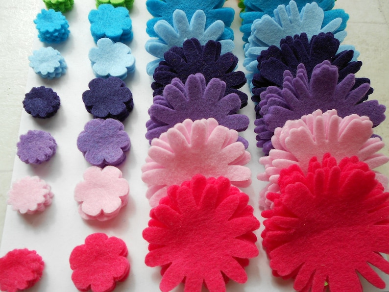 K&ouml;nnte beinhalten: Sortiment an Filzblumenformen in verschiedenen Farben, darunter Gr&uuml;n, Blau, Lila, Rosa und Rot. Die Blumen sind in Stapeln &uuml;bereinander gelegt und erzeugen einen Farbverlaufseffekt.