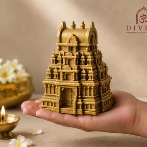 May include: A gold-coloured miniature temple statue featuring intricate architectural details, including a tiered gopuram-style roof and a central entrance. The text DIVINE HANDMADE STATUES HOME MANDIRS with an Om symbol is visible in the upper right corner.