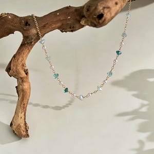 Könnte beinhalten: Eine zarte goldene Kette mit abwechselnd kleinen weißen Perlen und facettierten hellblauen sowie türkisfarbenen Kristallperlen. Die Kette ist über ein Stück Treibholz drapiert und zeigt ein schlichtes Gliederkettendesign mit gleichmäßig verteilten Edelsteinakzenten.