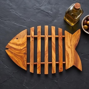 May include: A fish-shaped trivet crafted from light and dark brown wood with a visible grain pattern. The design features a head piece, a tail piece, and four vertical slats connected by two horizontal wooden rods, creating a skeleton-like structure.