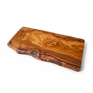 Puede incluir: Una tabla de servir rectangular de madera de olivo con un borde natural en uno de sus lados. La superficie muestra un patrón de veta rico y arremolinado en tonos marrón dorado y ámbar oscuro, con un pequeño orificio circular arriba a la izquierda.