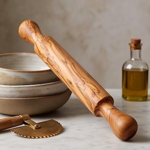 May include: A cylindrical wooden rolling pin with a distinct brown grain pattern, positioned diagonally. Nearby are two stacked ceramic bowls, a brass pastry cutter with a serrated edge, and a small glass bottle filled with golden oil.