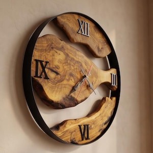 May include: A round wall clock featuring a black metal frame and natural wood segments with visible grain. The clock face displays black Roman numerals at the twelve, three, six, and nine o'clock positions, with metal hour, minute, and second hands.