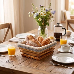 Op de afbeelding: Een vierkante houten broodmand met een lichtgekleurde stoffen voering met een blauwe patroonrand, gevuld met diverse soorten brood en croissants, geplaatst op een rustieke houten eettafel met borden, glazen sinaasappelsap en koffie.