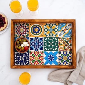 Puede incluir: Una bandeja de servicio rectangular de madera con una cuadrícula de doce azulejos de cerámica decorativos con varios patrones geométricos y florales coloridos. Un pequeño cuenco marrón con aceitunas verdes y cubos de queso blanco descansa sobre ella.