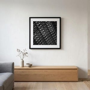 May include: A square black and white wall art print featuring a repeating geometric pattern of intersecting curved lines, encased in a thin black frame with a wide white mat border, displayed centered on a plain white wall above a wooden console.