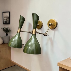 Puede incluir: Una lámpara de pared doble con dos pantallas cónicas de color verde oliva y detalles en latón. El aplique cuenta con brazos articulados ajustables conectados a dos soportes de pared circulares de latón, permitiendo un posicionamiento flexible.