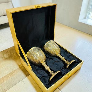 May include: Two gold-colored metal goblets with intricate embossed patterns on the bowls and stems, presented inside a rectangular gold-toned box lined with black fabric. The goblets feature a heart-shaped design element on the bowl surface.