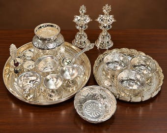 Può includere: Un set puja in metallo color argento con due lampade a olio decorate, un grande vassoio circolare con un vaso centrale, diverse ciotole piccole, un cucchiaio e una campanella. Un secondo vassoio contiene tre ciotole e una è posta in primo piano.