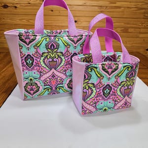 May include: Two tote bags of different sizes featuring a vibrant, multi-colored paisley and frog pattern on the front panels. The bags have solid light pink side panels and sturdy, bright purple woven fabric handles attached to the top.