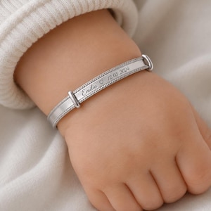 May include: A silver-toned metal identification bracelet featuring a flat, rectangular centre bar engraved with the name Emilia, a small heart symbol, and the date 12.05.2024. The bracelet has a rope-textured border and adjustable side links.