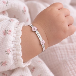 Może przedstawiać: Srebrzysta bransoletka typu bangle z dwoma małymi różowymi emaliowanymi kwiatkami z przezroczystymi kamieniami pośrodku. Metalowa opaska ma wygrawerowane imię Emma kursywą, umieszczone pomiędzy dwoma kwiatowymi akcentami na nadgarstku.