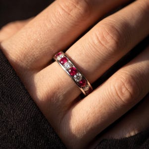 May include: A silver-toned band ring featuring an alternating channel-set pattern of round, vibrant red rubies and clear, sparkling white diamonds. The metal band has a smooth, polished finish that reflects light across the gemstones.
