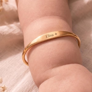 May include: A polished gold-tone metal bangle bracelet featuring a flat, centered bar engraved with the name Elisa followed by a small four-pointed star symbol. The bracelet has a smooth, rounded band design that encircles the wrist area.