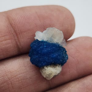 May include: A small mineral specimen featuring a vibrant, deep blue, textured cluster at its center. This central blue formation is flanked by a translucent, pale white crystalline structure on top and a rough, light beige mineral base at the bottom.