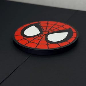 以下が含まれることがあります： スパイダーマンの象徴的な赤と黒のクモの巣模様が描かれた円形のコースター。鮮やかな赤の背景に黒いクモの巣が広がり、太い黒い縁取りで囲まれた2つの大きな白いアーモンド型の目がデザインされています。