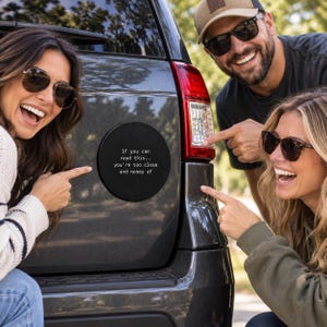Puede incluir: Un imán circular negro para coche con texto blanco que dice: If you can read this... you're too close and nosey af. El imán está colocado en el portón trasero de un vehículo gris oscuro, cerca del conjunto de luces traseras rojas y transparentes.