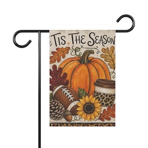 Könnte beinhalten: Eine rechteckige Gartenflagge mit dem Text 'Tis The Season' in schwarzer Schreibschrift. Die Illustration zeigt einen großen orangefarbenen Kürbis, einen braunen Football, eine Sonnenblume, einen Tannenzapfen, eine Eichel, Herbstblätter und einen Kaffeebecher mit Leopardenmuster-Manschette.