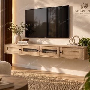 May include: A light oak floating media console featuring four cabinet doors, two of which have glass panels, and small black circular knobs. The unit is mounted horizontally on a wall beneath a large flat-screen television and decorative items.