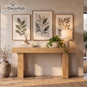 May include: A light wood console table sits against a wall featuring three framed botanical prints and the AHDecorHub logo. A ceramic vase with branches, stacked books, a potted plant, and a table lamp are arranged on the table surface.