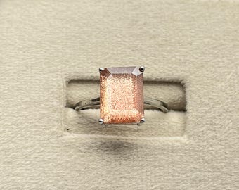 Könnte beinhalten: Ein Ring mit einem rechteckigen, facettierten Sonnenstein, der einen schimmernden, orange-braunen Glanz aufweist. Der Stein ist in eine schlichte, polierte Ringschiene aus silberfarbenem Metall gefasst und wird von vier Krappen gehalten.