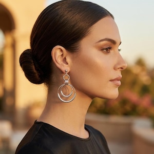 May include: A pair of silver-toned drop earrings featuring a small stud post connected to a series of three interlocking circular hoops. The two larger hoops have a textured, beaded surface, while the top hoop and post are smooth and polished.