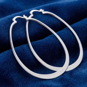 May include: A pair of silver-toned hoop earrings with an elongated, teardrop-shaped design. The earrings feature a smooth, polished metallic finish and a hinged closure mechanism at the top, creating a continuous, sleek, and minimalist oval silhouette.