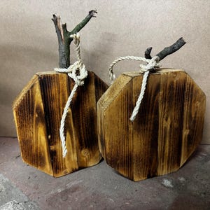 Pode incluir: Duas decorações de madeira em formato octogonal de abóbora com acabamento tingido escuro e veios de madeira visíveis. Cada peça é rematada com um caule de galho natural e acentuada com uma corda de juta torcida clara amarrada na base.