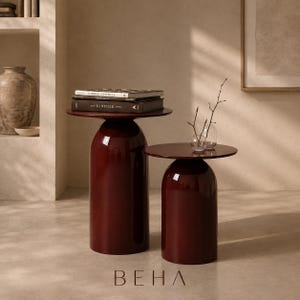May include: Two dark red, glossy, cylindrical side tables with flat circular tops. The taller table supports two stacked books titled Gabriel Chanel and The Kinfolk Home. The shorter table holds two small, clear glass vases with thin, bare branches.