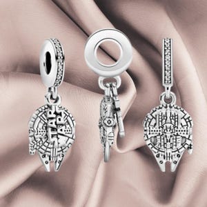 Może przedstawiać: Trzy srebrne zawieszki w kształcie statku kosmicznego Millennium Falcon. Dwie zawieszki pokazują widok z góry ze szczegółowymi detalami mechanicznymi i napisem STAR WARS, a środkowa zawieszka pokazuje widok z boku z okrągłym uchwytem.