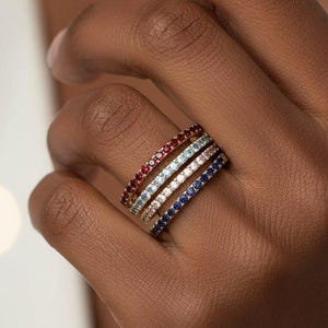 May include: A stack of four thin eternity bands worn on a finger. Each band features a continuous row of small, round-cut gemstones in distinct colours: deep red, light blue, clear white, and dark blue, set in a metallic band finish.