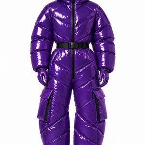 May include: A glossy purple puffer jumpsuit with a high collar, hood, and chevron-quilted pattern. It features a black front zipper, a black buckled waist belt, two large cargo pockets on the thighs, and matching purple gloves. The text PuffHaus is visible.