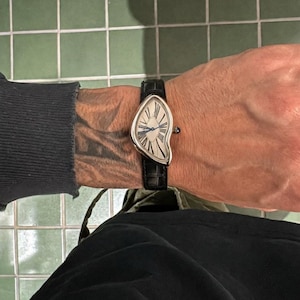 May include: A wristwatch with an asymmetrical, curved silver-tone case and a white dial featuring black Roman numerals and blue hands. The watch is attached to a black leather strap with a textured finish and includes a small blue crown detail.