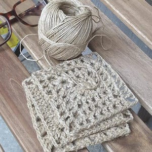 May include: A stack of crocheted squares made from natural jute twine, with a ball of the same twine and a pair of eyeglasses in the background.